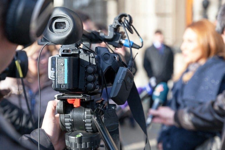 A television cameraman filming with a professional video camera at a press scrum, with a reporter holding a microphone toward an interviewee in the background