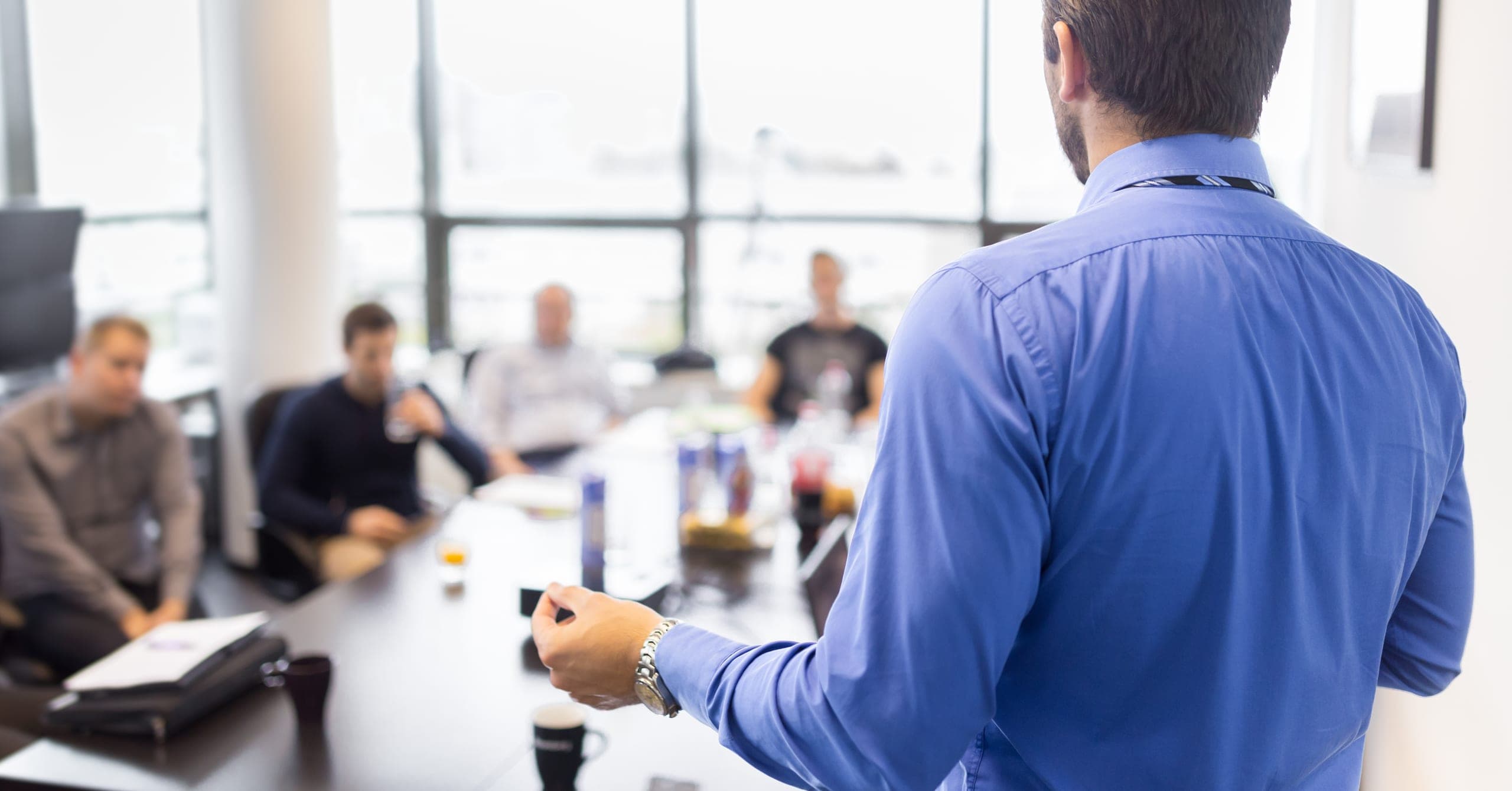 A man seen from behind in a blue shirt speaking and gesturing to colleagues seated around a conference table near floor-to-ceiling windows