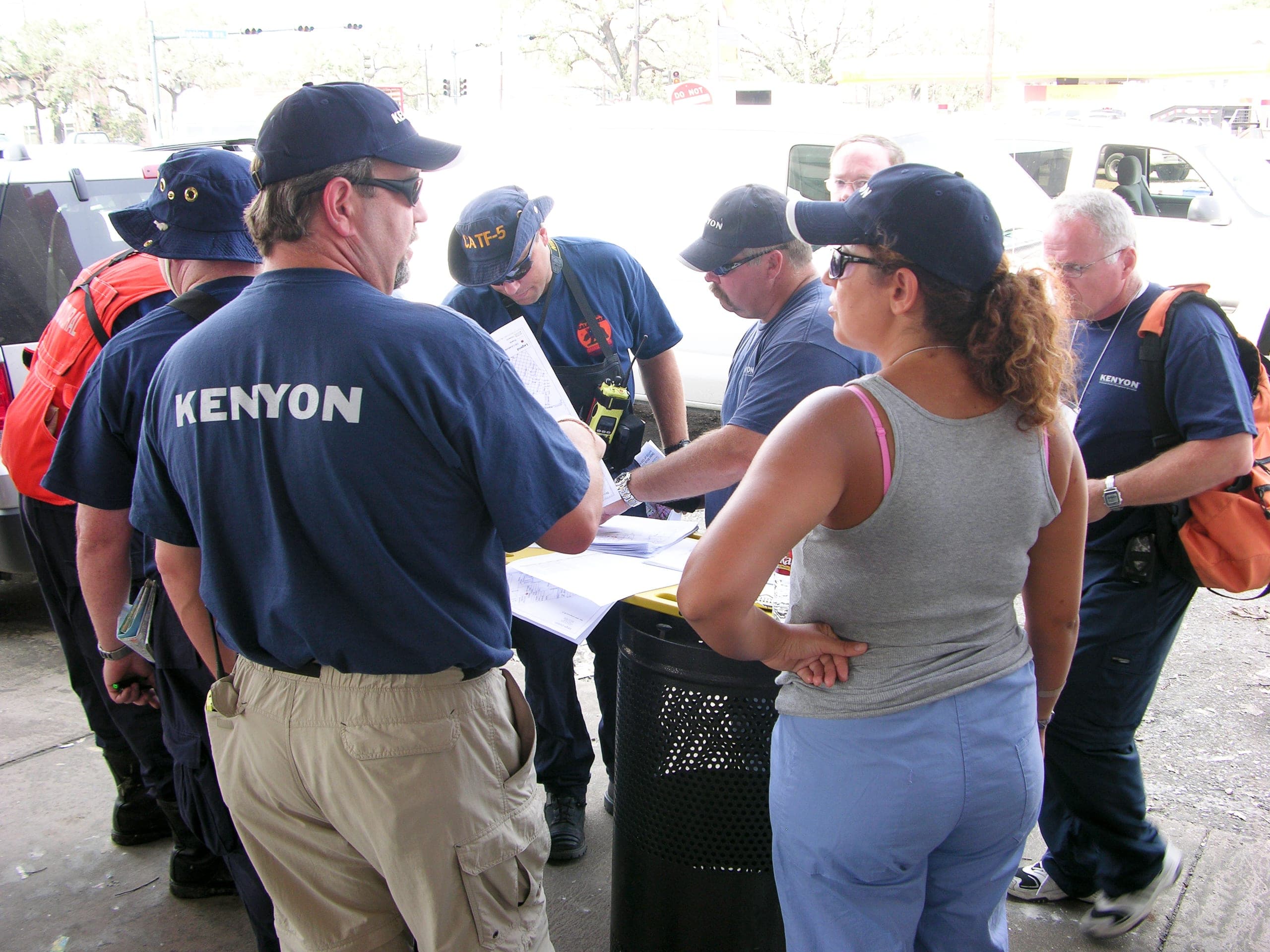 A group of responders in navy t-shirts and caps printed KENYON gathered around papers on a bin, talking with a woman in a grey tank top on an urban street