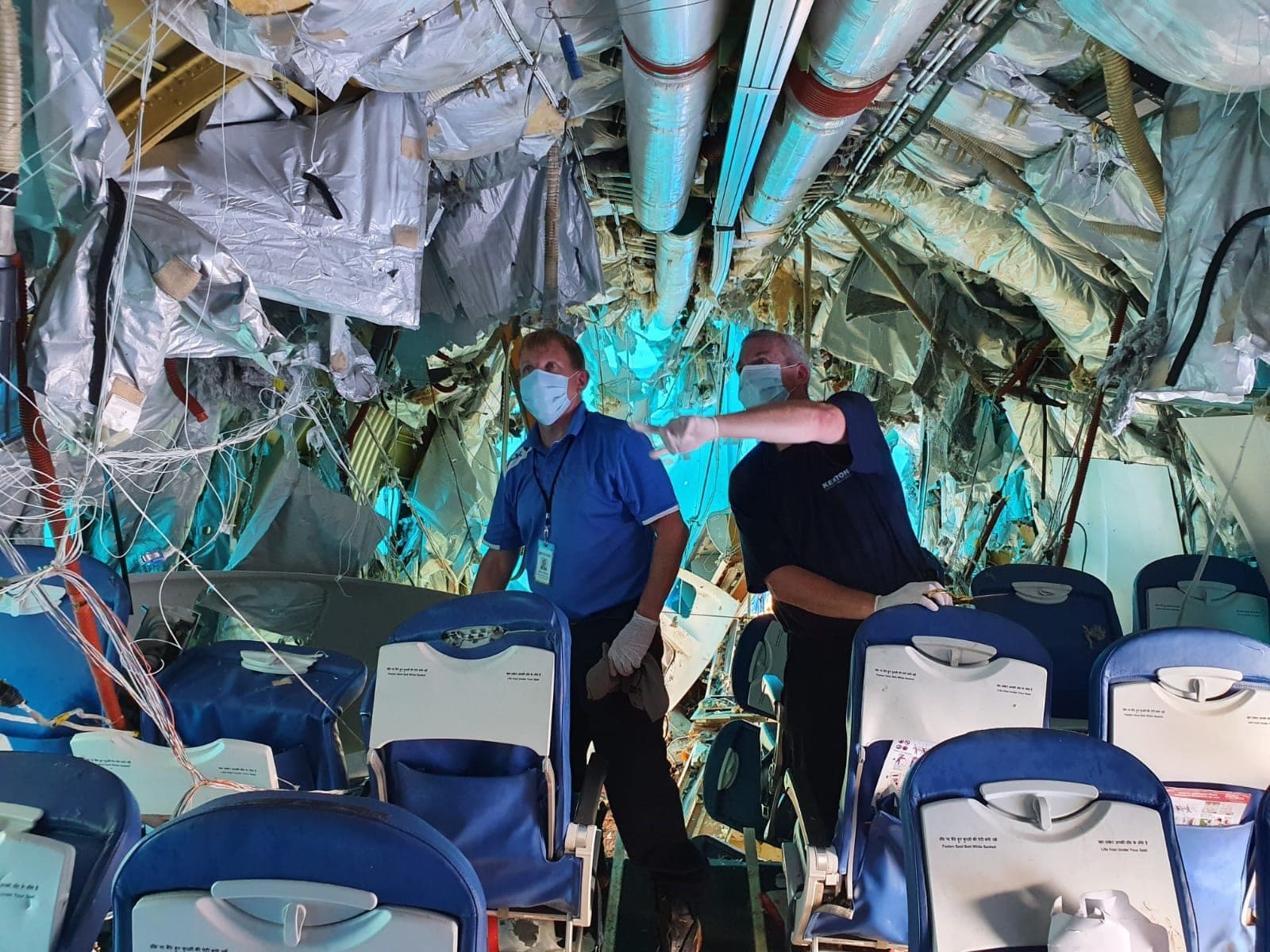Two men wearing surgical masks and gloves standing inside a heavily damaged aircraft cabin, with torn insulation and wreckage hanging overhead and rows of blue seats