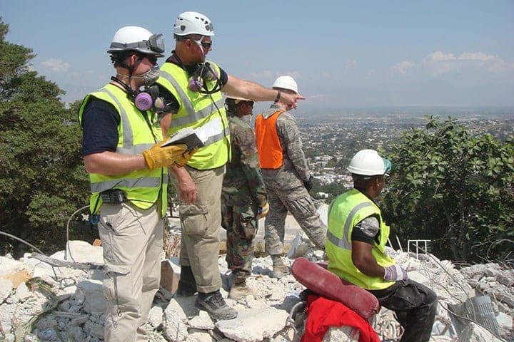 Responders in white helmets, yellow hi-vis vests and respirator masks standing on a hillside of collapsed concrete rubble, one holding a notebook and another pointing out towards the city spread across the valley below, with soldiers in camouflage behind them