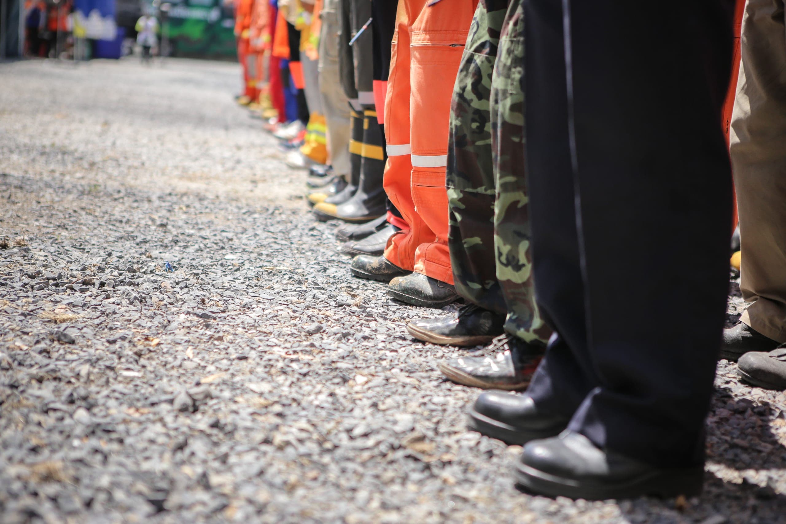 A line of people's legs and feet in a variety of uniforms, including orange coveralls and camouflage trousers, standing on gravel during a drill