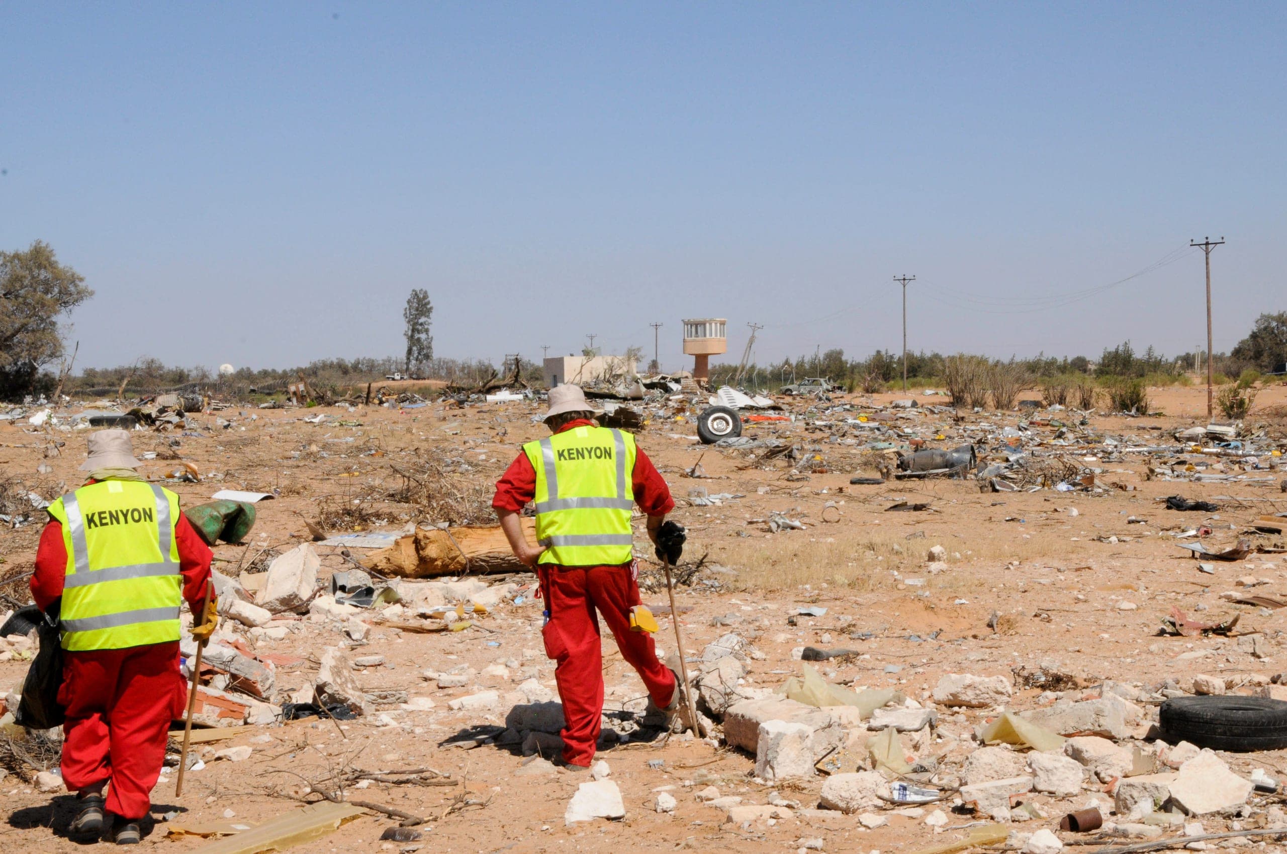 Two Kenyon responders in orange coveralls and yellow hi-vis vests printed with KENYON walking across an arid debris field, searching scattered wreckage and rubble, with a detached aircraft wheel and an airport tower in the distance