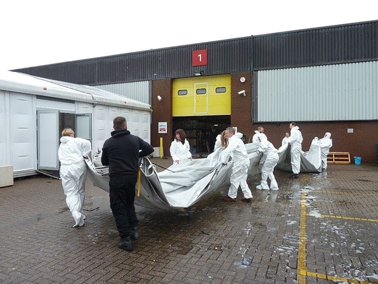 Workers in white protective coveralls carrying a large collapsed inflatable structure outside a warehouse marked with the number 1 and a yellow door
