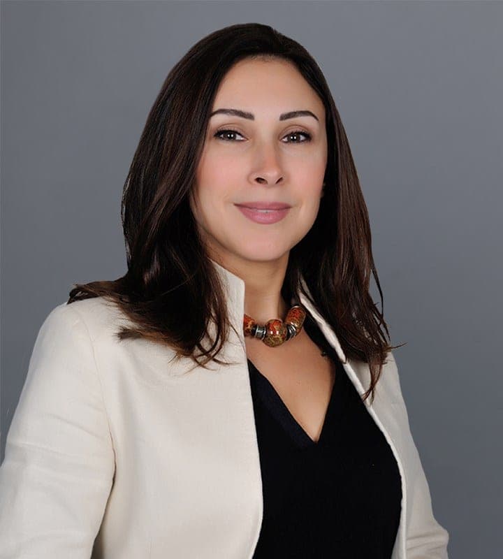 Portrait photo of a smiling woman with long dark hair, wearing a cream blazer over a black top with a multicolour beaded necklace, against a grey background