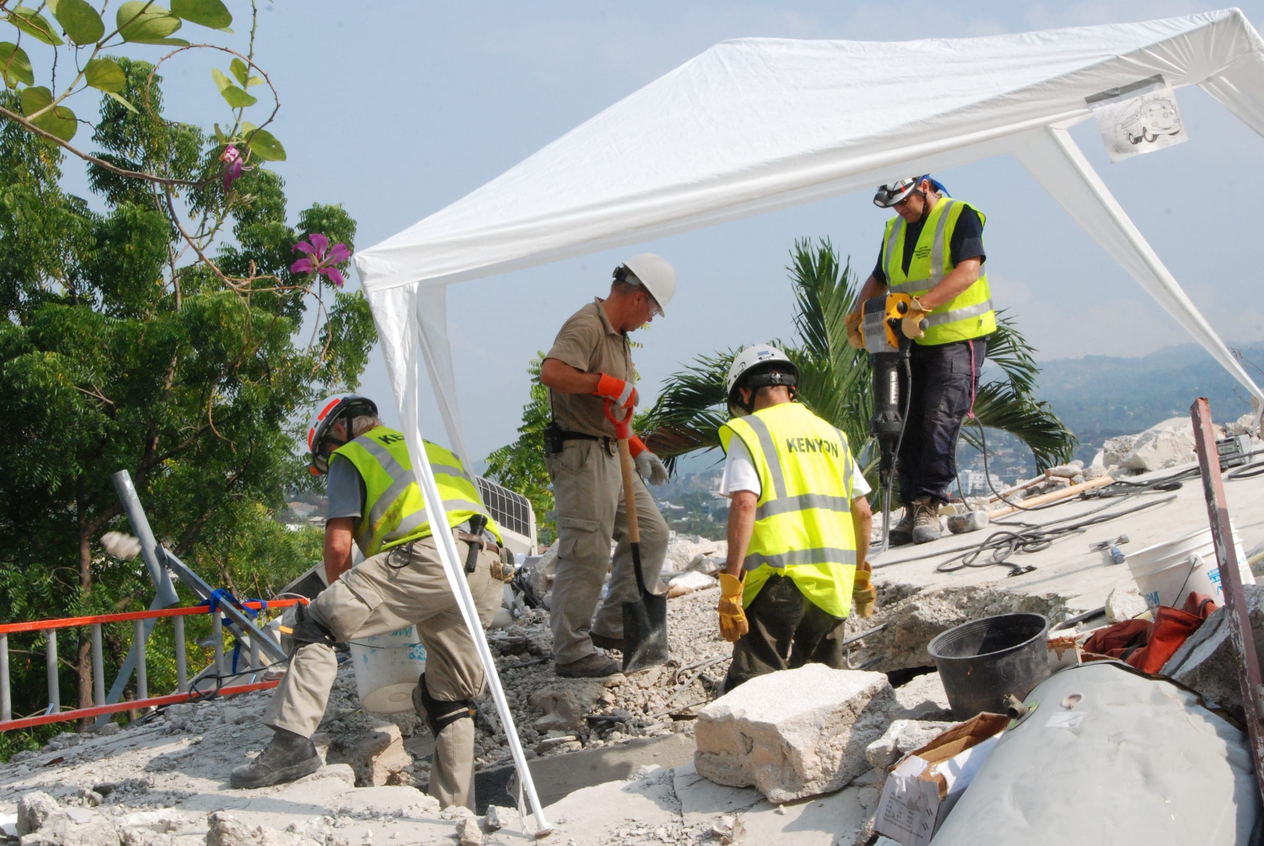 Responders in yellow hi-vis vests printed KENYON and white hard hats working under a white canopy tent on rubble, using a shovel and jackhammer, in a tropical setting