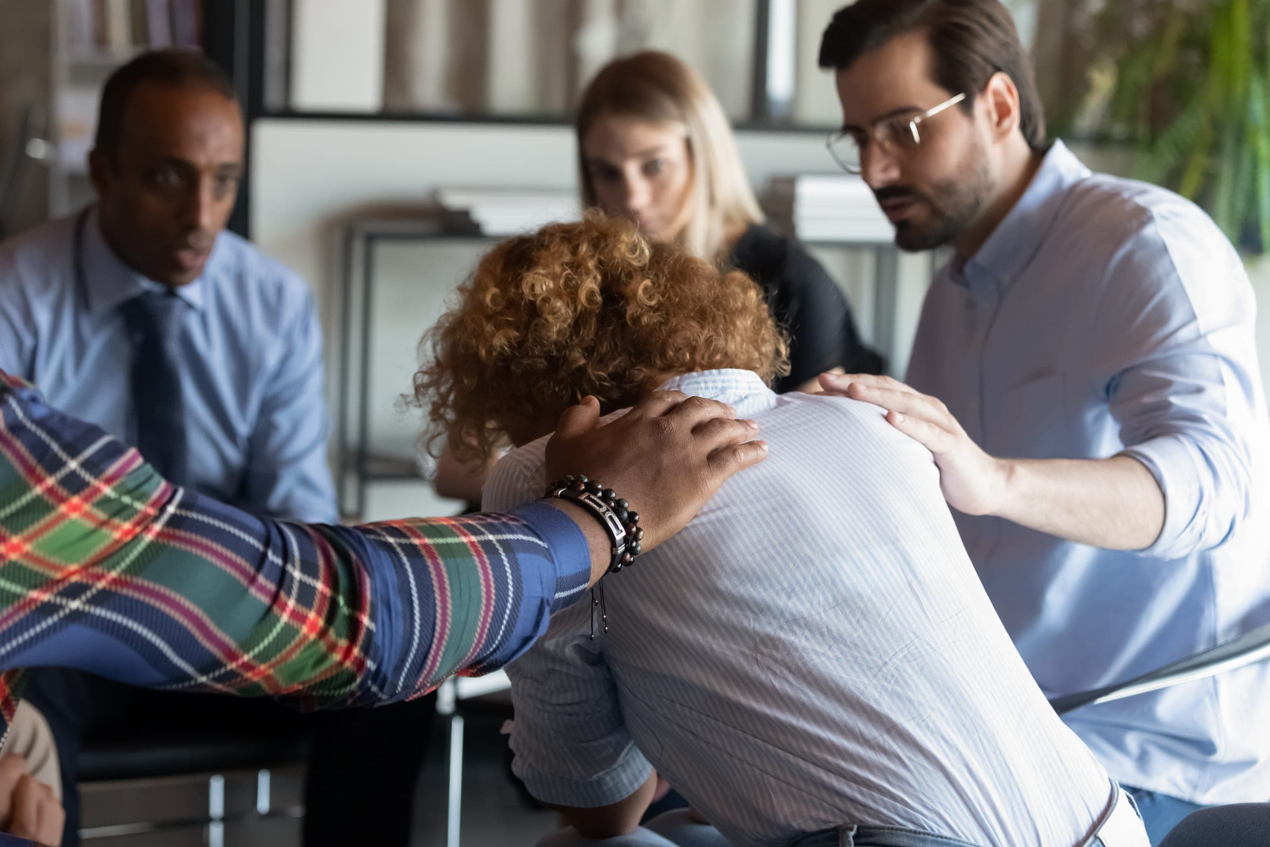 A group of colleagues gathered around a seated person, two of them resting a hand on the person's shoulders in comfort