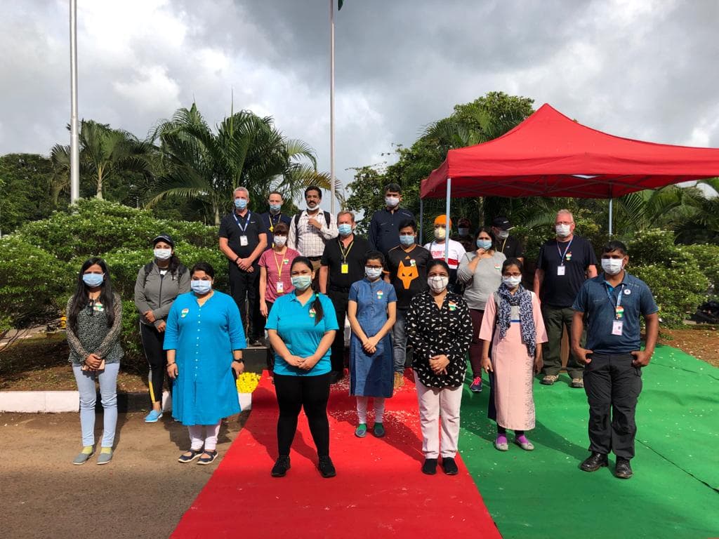 A group of about eighteen people wearing face masks standing on a red and green carpet at an outdoor ceremony, with a red canopy tent and palm trees behind them
