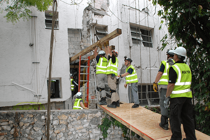 Kenyon responders in yellow hi-vis vests and white hard hats on a raised platform, lifting a wooden beam beside a damaged, partially collapsed white building
