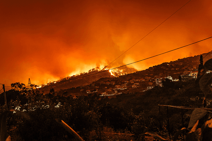 A wildfire glowing orange along a hillside ridge above a town at night, with smoke filling the sky and power lines in the foreground