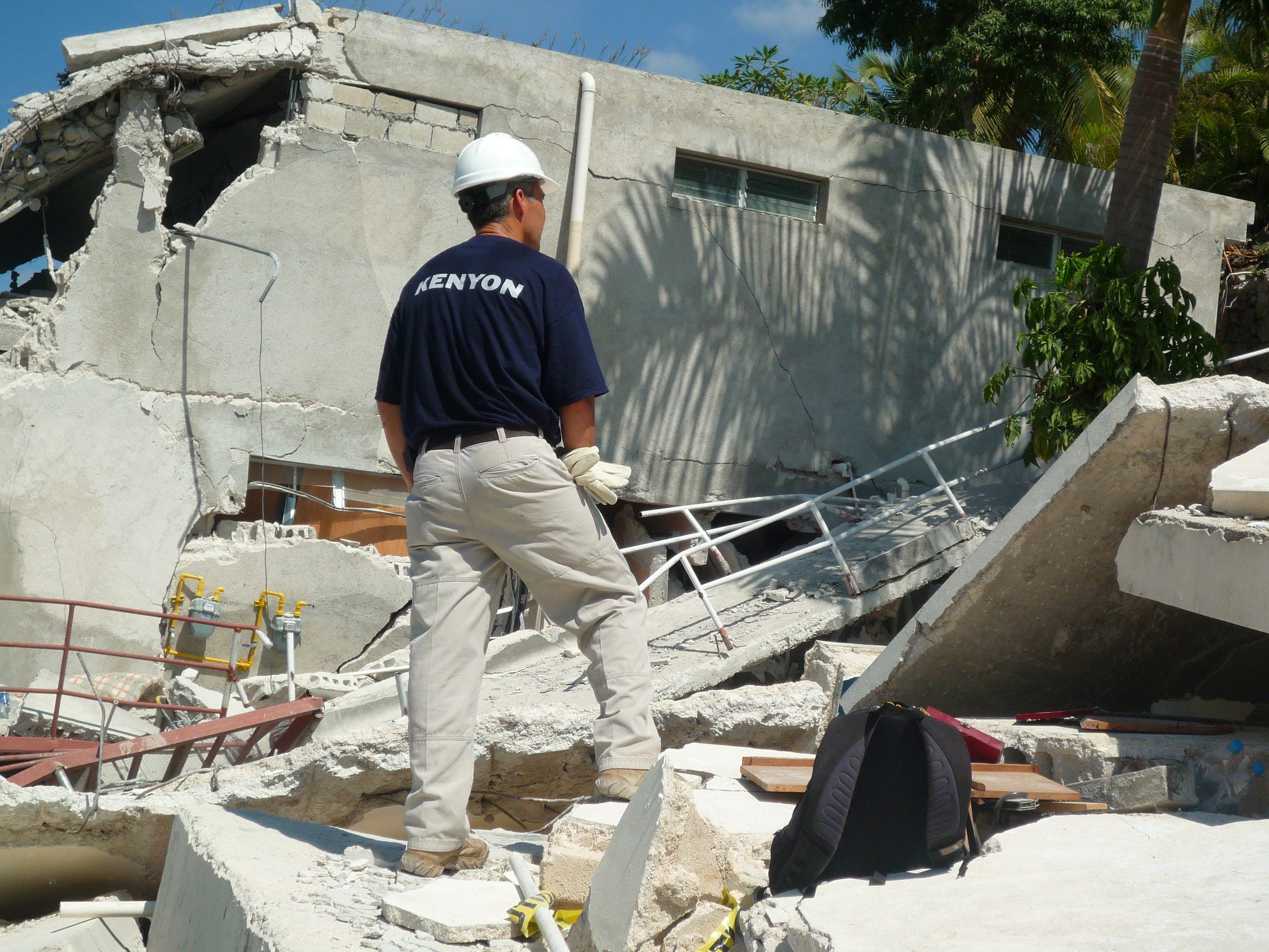 A responder wearing a navy KENYON t-shirt and white hard hat surveys earthquake rubble at a collapsed concrete building, with fallen railings, gas meters and debris around him