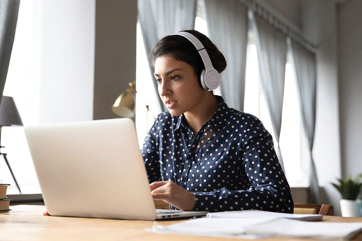 A woman wearing headphones working on a laptop at a desk near a window