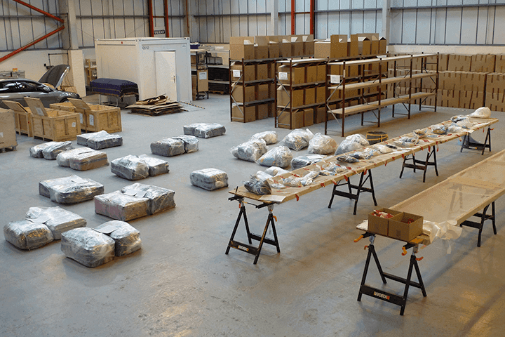 A warehouse with rows of shelving and wooden crates, personal belongings sealed in plastic bags laid out on trestle tables and the floor for processing