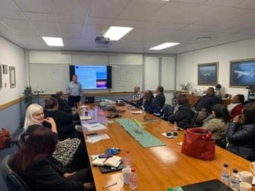 A training session in a meeting room, with a presenter standing at a screen and attendees seated around a table