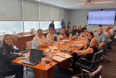 A group of people seated around a conference table in a boardroom, with a screen showing a spreadsheet in the background