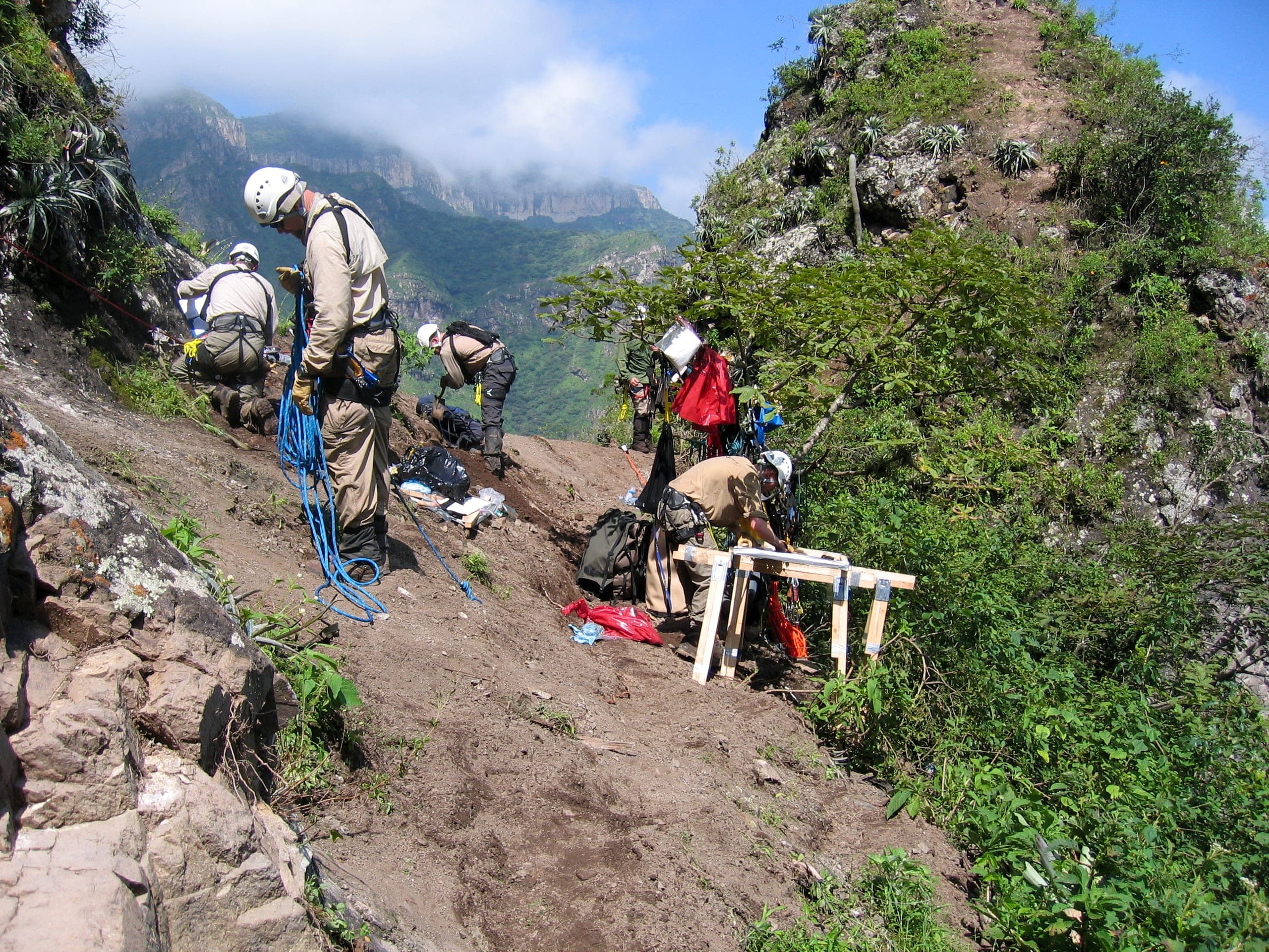 Responders in tan uniforms and white helmets with climbing ropes and harnesses working on a steep mountainside trail, with wooden equipment stands and mist-covered mountains behind them