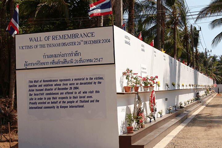 A long white memorial wall titled "Wall of Remembrance, Victims of the Tsunami Disaster 26th December 2004" with Thai flags and flowers placed along its base, palm trees behind