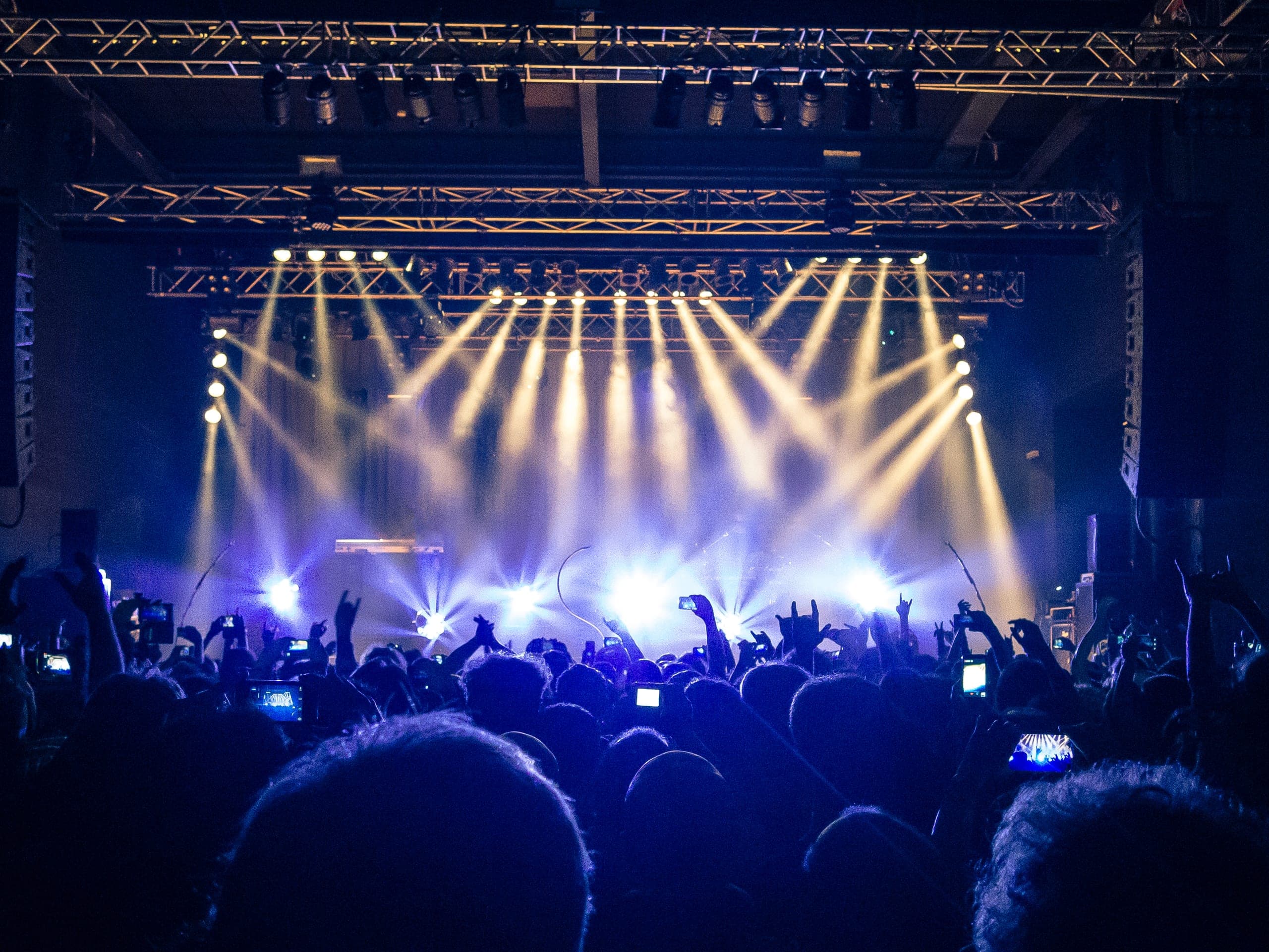 A concert crowd with hands raised and phones out, silhouetted against coloured stage lighting beams
