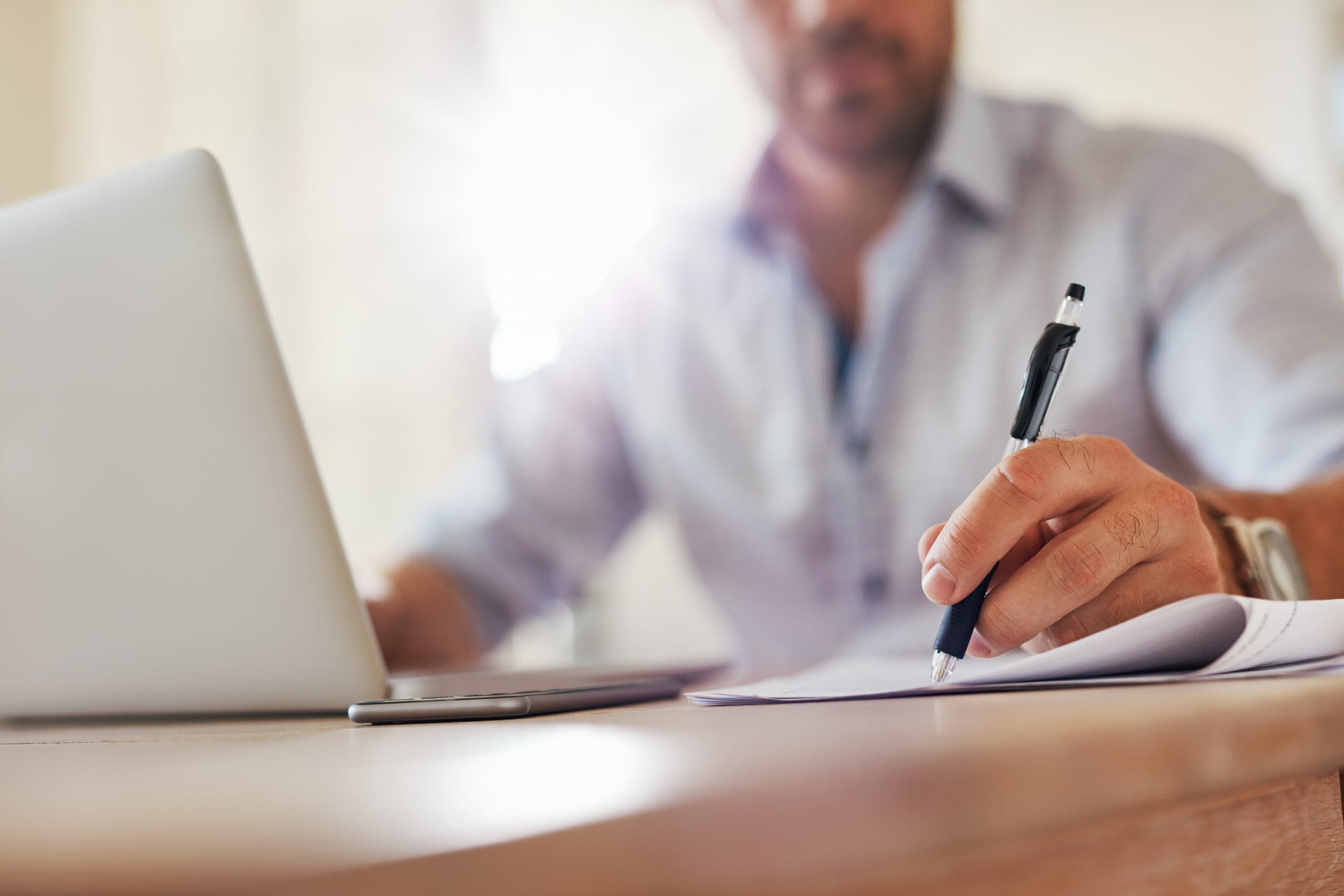 Close-up of a man's hand writing with a pen on paper beside an open laptop