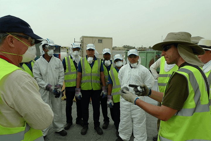 A responder briefing a line of workers in white protective coveralls, face masks and hi-vis vests, with a shipping container and vehicle in the background under a hazy sky