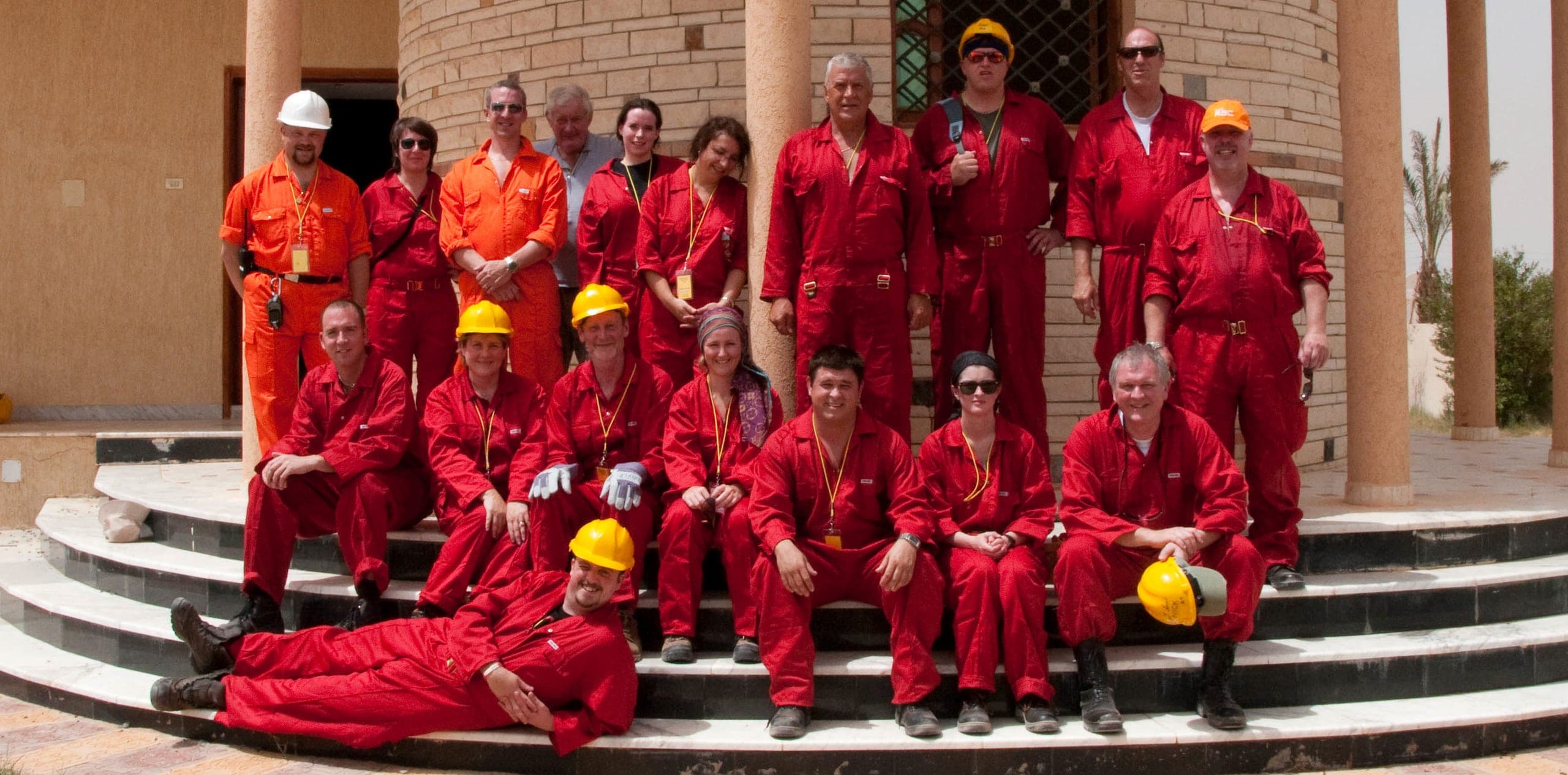 A group of about seventeen responders in red and orange coveralls, yellow hard hats and lanyards, posed on steps in front of an ornate colonnaded building