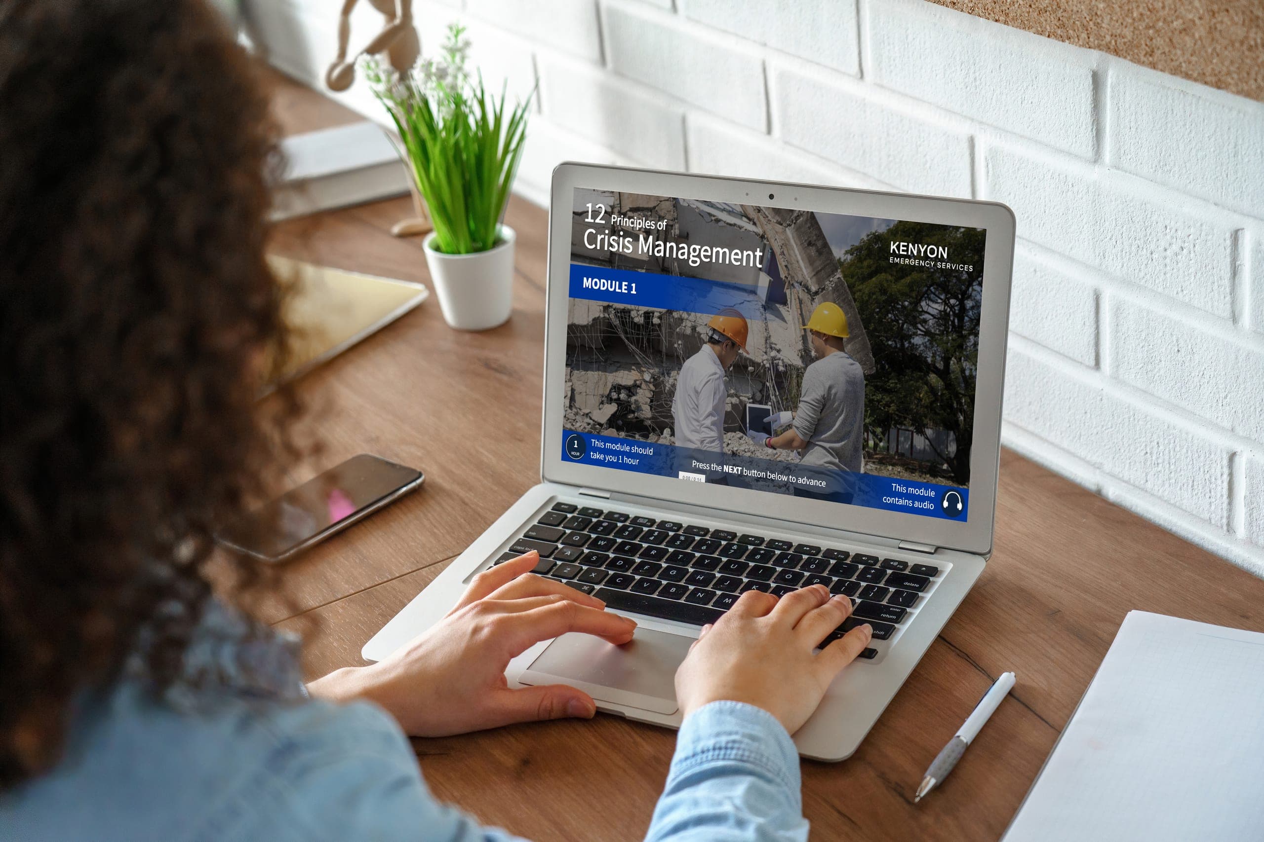 A laptop screen showing a Kenyon Emergency Services e-learning module titled "12 Principles of Crisis Management, Module 1" with a photo of two responders in hard hats inspecting rubble, a woman's hands typing on the keyboard